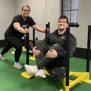 Two people exercising in a gym, one pushing a sled while the other sits on it.