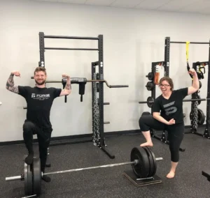 Two people flexing muscles beside a barbell in a gym setting.